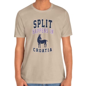 Può includere: T-shirt beige con la scritta "SPLIT HAPPENS IN CROATIA" in blu navy e viola. Una grafica di asino blu navy è centrata sotto il testo. La maglietta è a maniche corte e sembra essere di cotone.