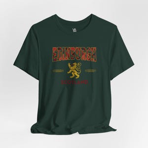 Puede incluir: Camiseta verde oscuro con la palabra "EDINBURGH" en un dise&ntilde;o de cuadros, un emblema de le&oacute;n dorado y la palabra "SCOTLAND" en rojo. La camiseta tiene mangas cortas y cuello redondo.