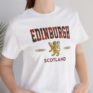 Puede incluir: Camiseta blanca con la palabra "EDINBURGH" en un dise&ntilde;o de cuadros rojos y verdes. Debajo, un emblema de le&oacute;n dorado y la palabra "SCOTLAND". La camiseta tiene cuello redondo y mangas cortas.
