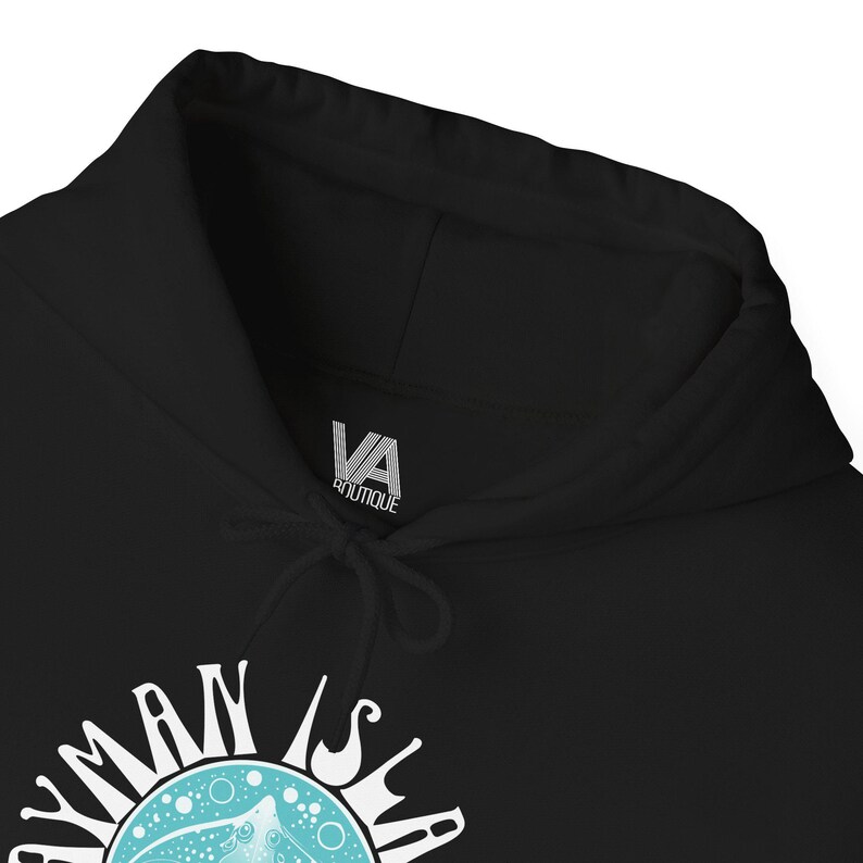 May include: Black hooded sweatshirt with a drawstring and the text "CAYMAN ISLANDS" in white lettering. The design includes a turquoise circle with a sea creature illustration. The brand logo "VA Boutique" is visible on the hood.
