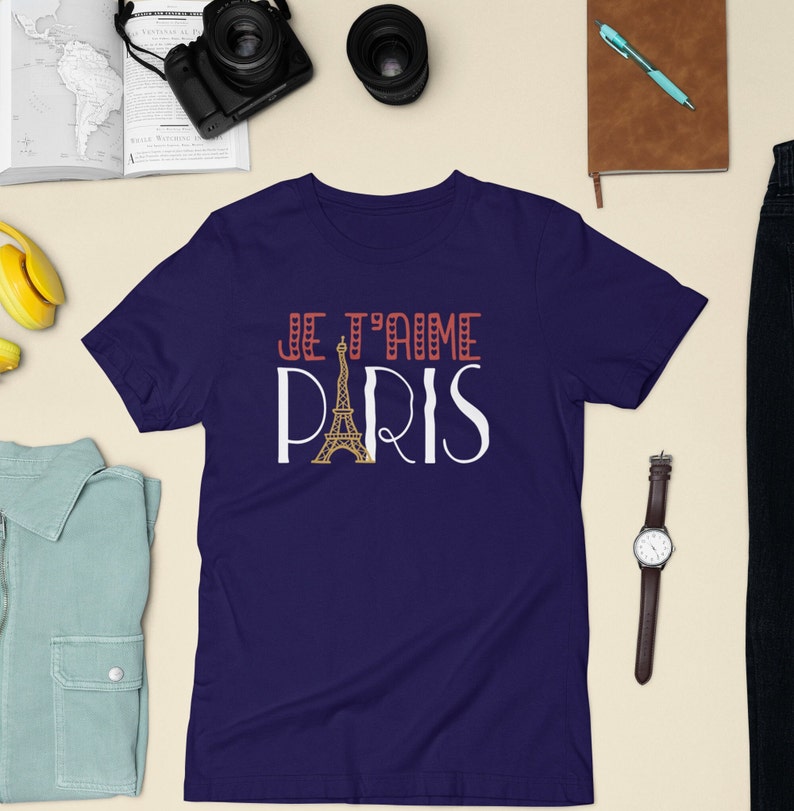 A tshirt that says "Je T'aime Paris" surrounded by travel gear. The A in "Paris" is replaced with a graphic of the Eiffel Tower.