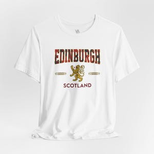 Puede incluir: Camiseta blanca con la palabra "EDINBURGH" en un dise&ntilde;o escoc&eacute;s, un emblema de le&oacute;n dorado y la palabra "SCOTLAND" en borgo&ntilde;a. La camiseta tiene mangas cortas y cuello redondo.