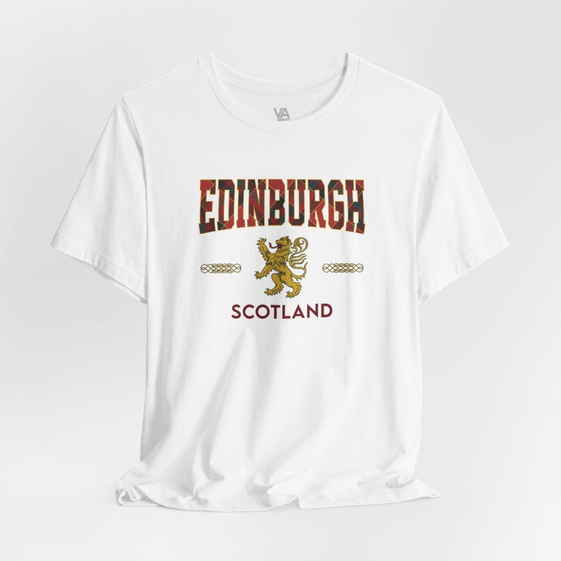 Puede incluir: Camiseta blanca con la palabra "EDINBURGH" en un dise&ntilde;o escoc&eacute;s, un emblema de le&oacute;n dorado y la palabra "SCOTLAND" en borgo&ntilde;a. La camiseta tiene mangas cortas y cuello redondo.