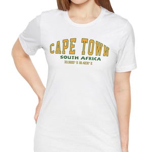 May include: White t-shirt featuring the text "CAPE TOWN" in gold and green. Below the text are the words "SOUTH AFRICA" and coordinates. The shirt has a crew neck and short sleeves.
