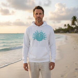 May include: White hoodie with a front pocket and a light blue graphic that reads "CAYMAN ISLANDS" around a stylized stingray. The hoodie is worn on a beach with the ocean in the background.