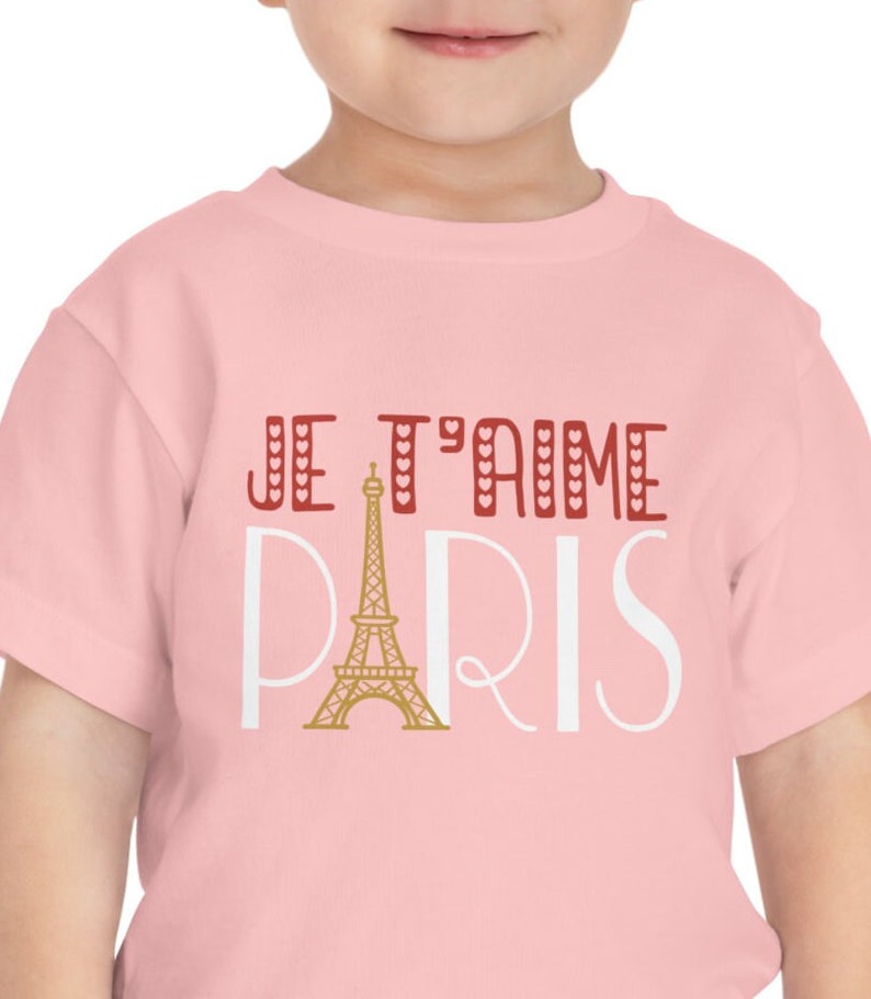 A todder wearing a pink tshirt that says "Je T'aime Paris". The A in "Paris" is replaced with a graphic of the Eiffel Tower.