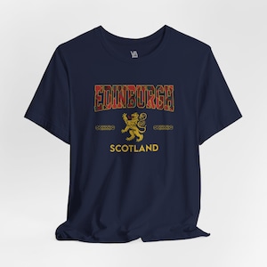 Puede incluir: Camiseta azul marino con la palabra "EDINBURGH" en un dise&ntilde;o escoc&eacute;s. Un emblema de le&oacute;n dorado y la palabra "SCOTLAND" est&aacute;n debajo. La camiseta tiene mangas cortas y cuello redondo.