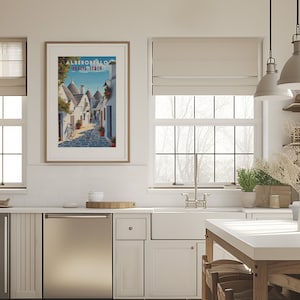 May include: A kitchen with white cabinets, a stainless steel dishwasher, and a white farmhouse sink. A framed poster with the text "ALBEROBELLO PUGLIA, ITALY" hangs on the wall above the sink. The poster features a colorful illustration of a street in Alberobello, Italy, with traditional trulli houses.