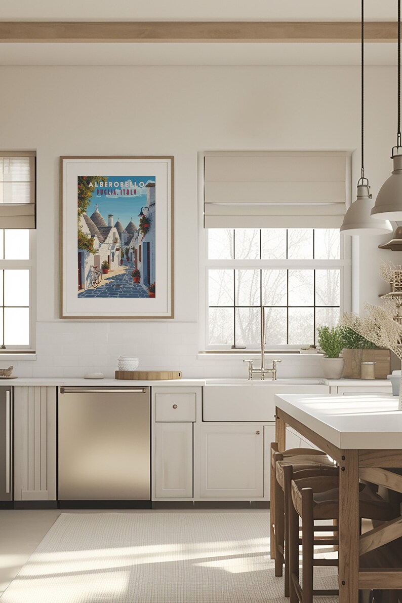 May include: A kitchen with white cabinets, a stainless steel dishwasher, and a white farmhouse sink. A framed poster with the text "ALBEROBELLO PUGLIA, ITALY" hangs on the wall above the sink. The poster features a colorful illustration of a street in Alberobello, Italy, with traditional trulli houses.