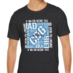 Könnte beinhalten: Dunkelgraues T-Shirt mit weißem und blauem Grafikdesign. Das Design zeigt Flughafen-Codes wie FRA, JFK und IAD, angeordnet in einem kreisförmigen Muster mit dem Text "If you can decode this, you speak my language."