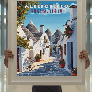 May include: A vintage travel poster featuring a colorful illustration of a street in Alberobello, Puglia, Italy. The street is lined with white buildings with conical roofs, and there are potted plants and a bicycle in the foreground.