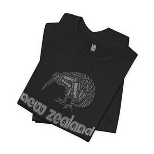 Puede incluir: Camiseta negra con un contorno blanco de un kiwi formado por palabras relacionadas con Nueva Zelanda. Las palabras "new zealand" est&aacute;n escritas en una fuente estilizada debajo del p&aacute;jaro.
