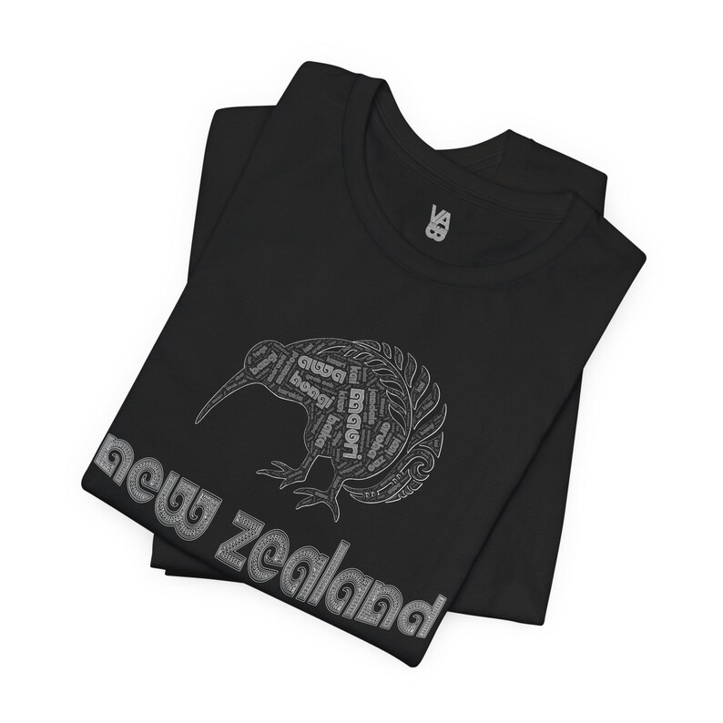 Puede incluir: Camiseta negra con un contorno blanco de un kiwi formado por palabras relacionadas con Nueva Zelanda. Las palabras "new zealand" est&aacute;n escritas en una fuente estilizada debajo del p&aacute;jaro.