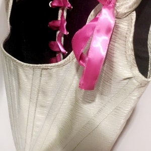 May include: A white corset with pink satin ribbon lacing. The corset has a black lining and is made of a textured fabric.