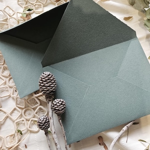 Op de afbeelding: Twee open, saliegroene enveloppen met een getextureerde afwerking. De binnenkant van een envelop is donkergroen. Drie dennenappels op slanke stelen staan op de voorgrond, met een witte, getextureerde achtergrond.