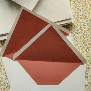 Op de afbeelding: Een stapel open enveloppen met een helderwitte buitenkant en een getextureerde binnenvoering in een diepe terracottakleur. De enveloppen hebben een klassiek driehoekig klepontwerp, wat het contrast benadrukt tussen het witte papier en de warme, aardse rood-oranje kleur aan de binnenzijde van het briefpapier.