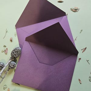 Op de afbeelding: Twee open enveloppen in een diepe pruimpaarse kleur met een gladde, matte textuur. De enveloppen hebben een klassiek ontwerp met een puntige klep en liggen overlappend over elkaar, waardoor de binnen- en buitenoppervlakken zichtbaar zijn.