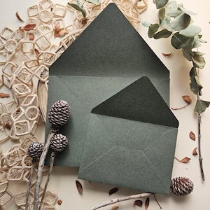 Op de afbeelding: Twee open enveloppen in een donkere bosgroene kleur met een textuurafwerking. De enveloppen worden getoond met een tak met kleine dennenappels en gedroogde eucalyptusbladeren, gerangschikt naast decoratieve crèmekleurige macramé-elementen.