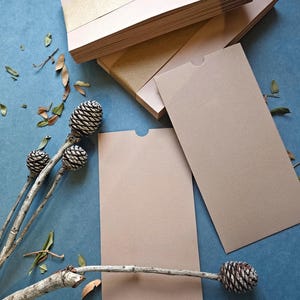 Può includere: Un mucchio di biglietti di carta beige con una fascia dorata, insieme a biglietti singoli e una busta abbinata. I biglietti sono disposti su una superficie blu scuro, con pigne e rametti decorativi.