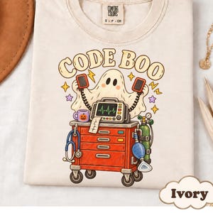 May include: An ivory t-shirt featuring a graphic of a ghost holding defibrillator paddles above a red medical crash cart. The cart includes a monitor, a blue stethoscope, a green oxygen tank, and a purple mug with a jack-o'-lantern face. The text reads CODE BOO.