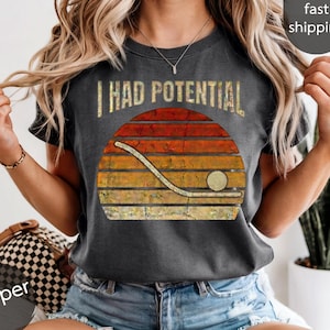 Puede incluir: Camiseta gris oscuro con el texto "I HAD POTENTIAL" sobre un diseño de palo y disco de hockey sobre un degradado de puesta de sol. La camiseta tiene un aspecto vintage y desgastado.