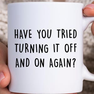 Könnte beinhalten: Weiße Keramiktasse mit dem schwarzen Text "HAVE YOU TRIED TURNING IT OFF AND ON AGAIN?". Die Tasse wird von einer Person gehalten und hat einen gebogenen Griff. Standardgröße.