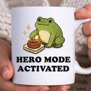 以下が含まれることがあります： 白いセラミックマグカップ。漫画の緑色のカエルが「HERO」ボタンを押しています。「HERO MODE ACTIVATED」の文字が黒で書かれています。面白くてユニークなアイテムです。