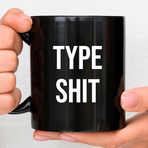 May include: A black ceramic mug with the words "TYPE SHIT" printed in large, white capital letters. The mug has a black handle and is held by a person.