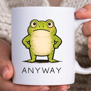 May include: White ceramic mug featuring a cartoon frog with a grumpy expression. The frog is green with yellow accents and is standing with its arms crossed. The word "ANYWAY" is printed in black below the frog.