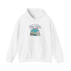 May include: A white hooded sweatshirt featuring a central graphic of three stacked books topped with blue ocean waves and a small orange sun. The text "LOST BETWEEN PAGES & WAVES" is printed above the illustration, which also includes seashells and a starfish.