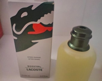 May include: A green and white aftershave bottle with a green alligator logo and the text "Booster Lacoste" on the label. The bottle is next to a white box with the same logo and text.