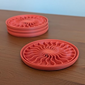May include: A set of four coral-colored, round coasters with a spiral design. One coaster is separate, while the other three are stacked. The coasters are made of a solid material and sit on a wooden surface.
