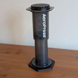May include: AeroPress coffee maker, a dark gray cylindrical device with the brand name printed in white. The device has a black hexagonal base and a black top with a ridged surface. The coffee maker is on a wooden surface.