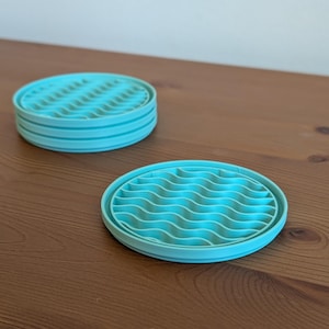 May include: Two turquoise silicone coasters with a wavy design are stacked on a wooden surface. One coaster is separate, showing the wavy pattern. The coasters are round and have a ridged surface.