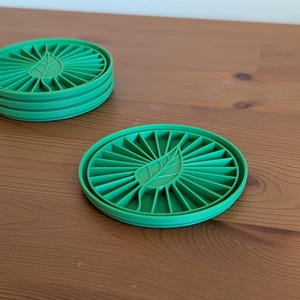May include: A stack of four and a single green, circular coasters with a leaf design in the center. The coasters are made of a ridged material and sit on a wooden surface.