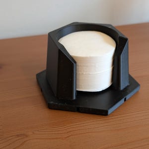 May include: A black, geometric coffee filter holder with a stack of white paper filters inside. The holder has a hexagonal base and a matching top section with cutouts. The holder is sitting on a wooden surface.