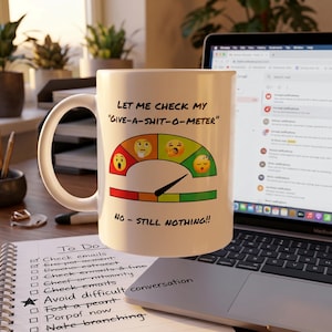Könnte beinhalten: Weiße Keramiktasse mit dem Text "LET ME CHECK MY GIVE-A-SHIT-O-METER" und "NO - STILL NOTHING!!". Die Tasse zeigt ein Messgerät mit Emoji-Gesichtern. Eine To-Do-Liste und ein Laptop sind im Hintergrund.