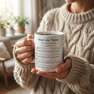 Pode incluir: Uma caneca de cerâmica branca com o "Tema do Lago dos Cisnes" de Piotr Ilitch Tchaikovsky. A caneca, segurada por uma pessoa com um suéter bege de malha de cabo, exibe notas musicais e texto. Vapor sobe da caneca.