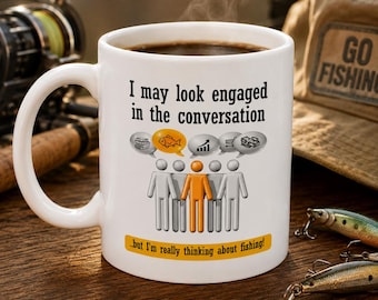 May include: A white ceramic mug featuring the black text I may look engaged in the conversation, a graphic of stick figures with speech bubbles containing icons, and the orange text ...but I'm really thinking about fishing! on a yellow banner.