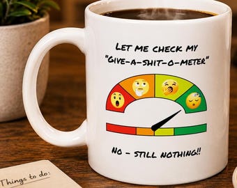 May include: White ceramic coffee mug with black text that reads "Let me check my 'Give-a-shit-o-meter'" and "No - still nothing!!" The mug features a gauge with various emoji expressions.