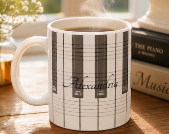 May include: A white ceramic mug featuring a black and white piano key pattern overlaid with horizontal musical staff lines. The name Alexandria is printed in a black cursive script across the center of the mug, which contains a dark liquid.
