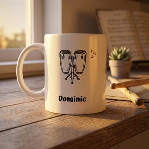 Puede incluir: Taza de cerámica blanca con un dibujo negro de dos congas sobre un soporte, con notas musicales. El nombre "Dominic" está impreso debajo. Una planta suculenta y baquetas están en el fondo.