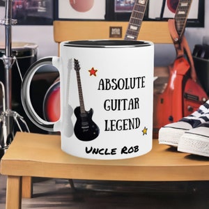 May include: White ceramic mug with a black interior and handle. The mug features a black guitar graphic and the text "ABSOLUTE GUITAR LEGEND UNCLE ROB" with star accents. The mug is on a wooden surface.