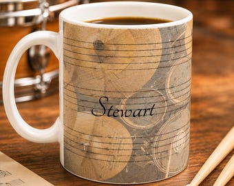 May include: A white ceramic mug featuring a graphic print of metallic drum cymbals overlaid with black musical staff lines. The name Stewart is printed in a black cursive font across the center of the mug, which has a handle on the left side.