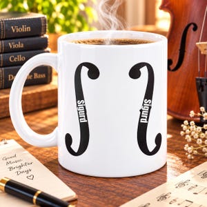 May include: A white ceramic mug featuring two black f-hole shapes from a string instrument, each printed with the name Sigurd in white vertical text. The mug is filled with a dark steaming beverage and has a curved handle on the left side.