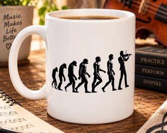 May include: A white ceramic mug featuring a black graphic print that depicts the evolution of humans, starting from a primate and progressing to a person playing a violin. The mug is filled with a dark liquid and has a smooth, glossy finish.