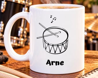 May include: A white ceramic mug featuring a black line drawing of a snare drum with two drumsticks resting on top and musical notes above. The name Arne is printed in black text below the drum illustration. The mug contains a hot beverage.
