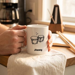 Puede incluir: Taza de cerámica blanca con un dibujo lineal negro de dos bongós y notas musicales. El nombre "Jimmy" está impreso debajo de los bongós. La taza está sostenida por una persona.