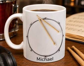 May include: A white ceramic mug featuring a graphic of a snare drum with two wooden drumsticks resting on top. The name Michael is printed in black text at the bottom of the drum graphic. The mug contains a dark liquid with steam rising from the top.