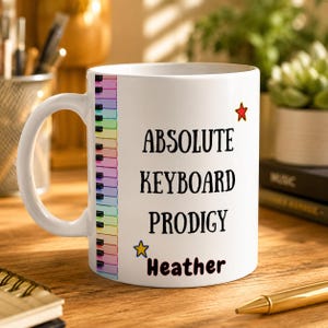 Może przedstawiać: Biały ceramiczny kubek z motywem klawiatury fortepianu. Kubek zawiera napis "ABSOLUTE KEYBOARD PRODIGY" z imieniem i gwiazdkami. Idealny prezent dla muzyka.